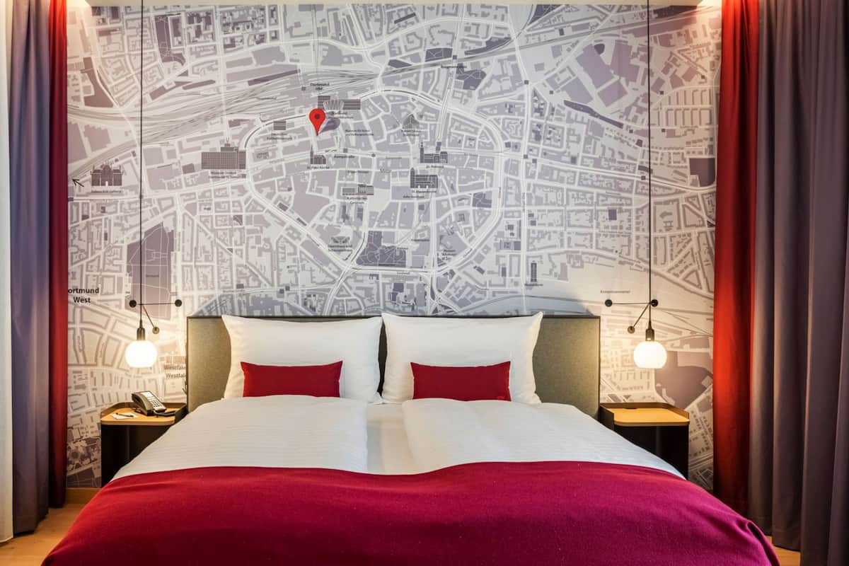 A hotel room with a large bed, red bedding, a map wallpaper, and two bedside lamps.