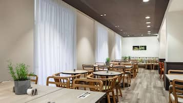 En modern restaurang med träbord och stolar, stora fönster med vita gardiner och en platt-TV på väggen.