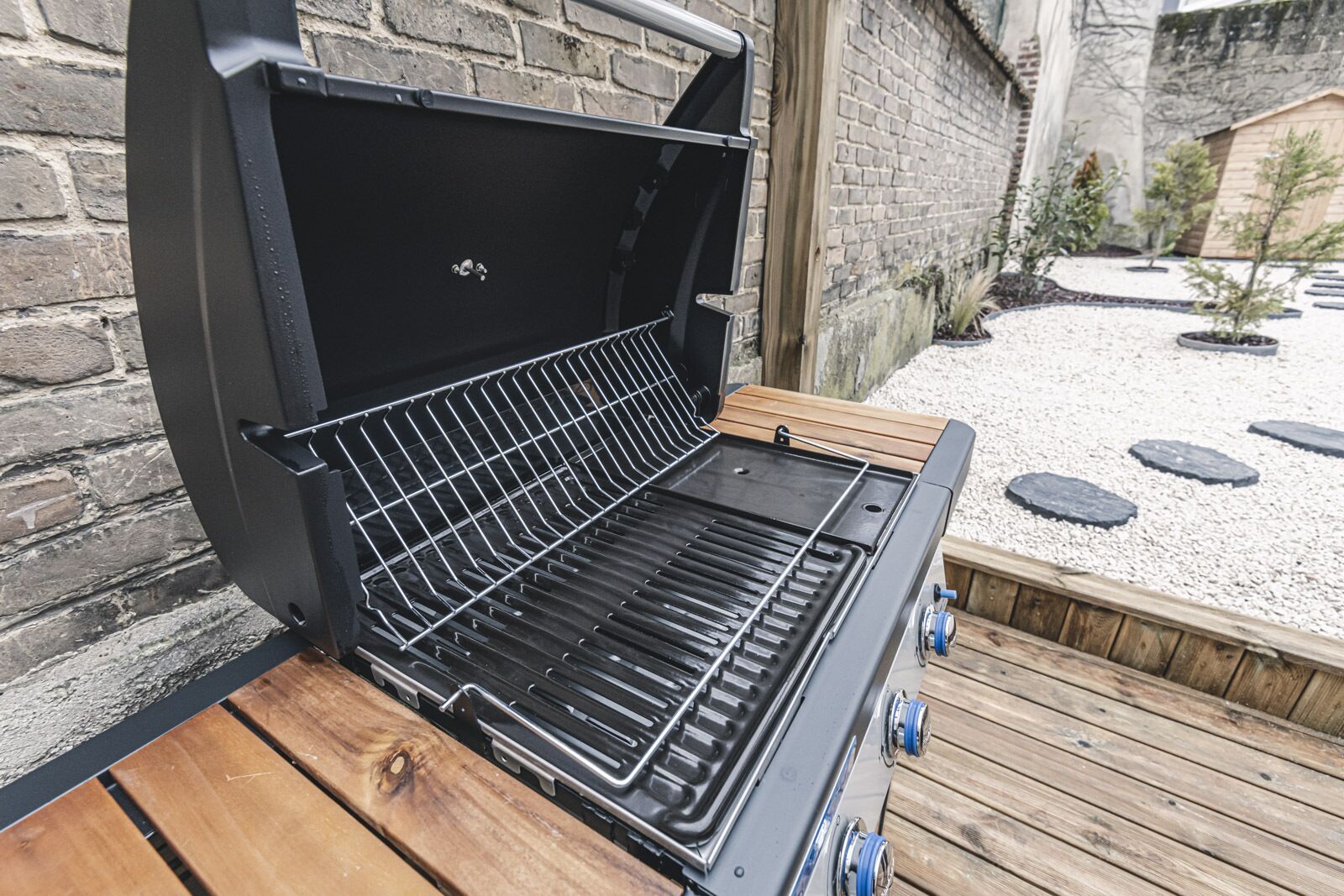 A barbecue grill with a metal rack, set on a wooden deck with a brick wall in the background.