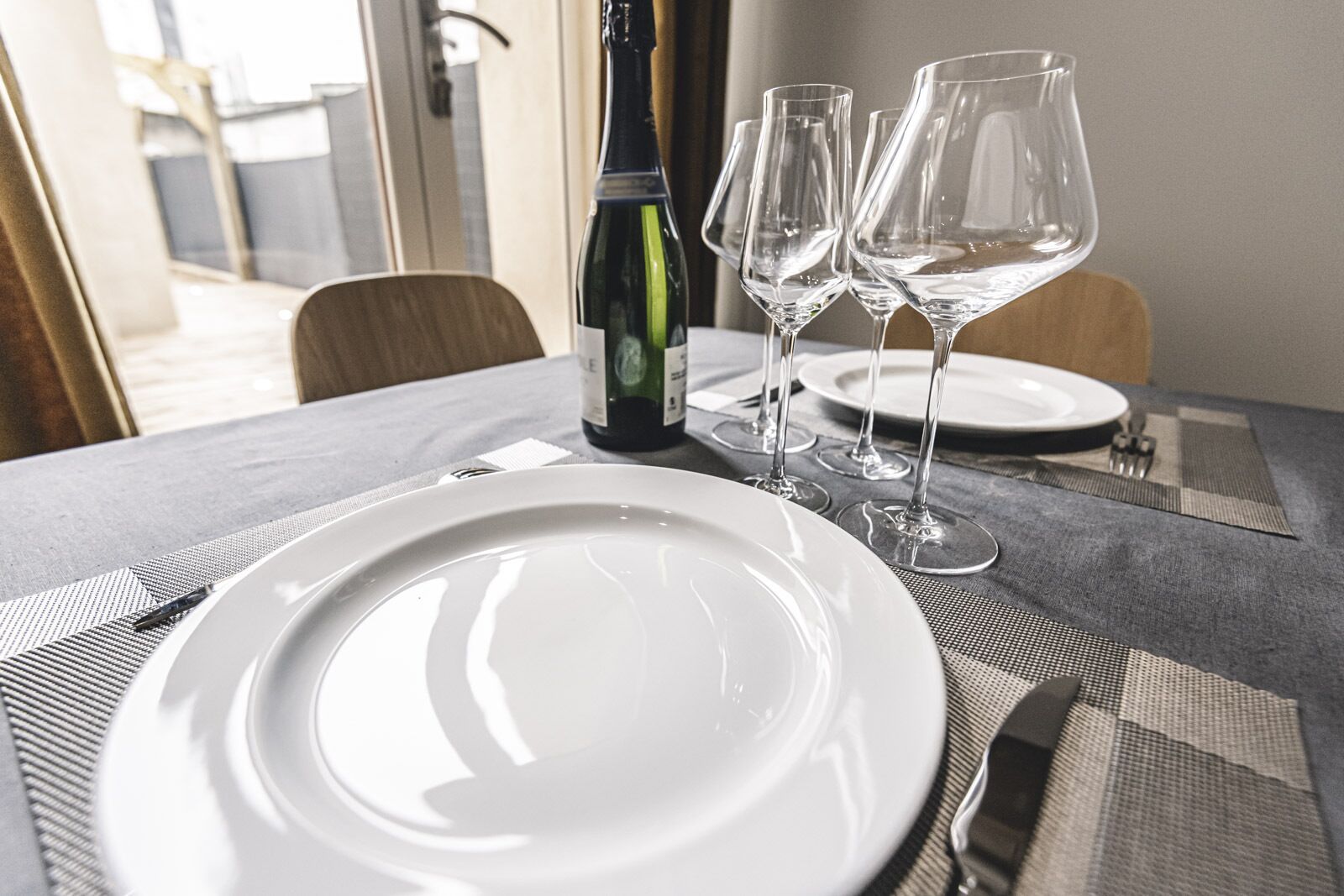 A dining table set with a white plate, two wine glasses, and a bottle of wine.