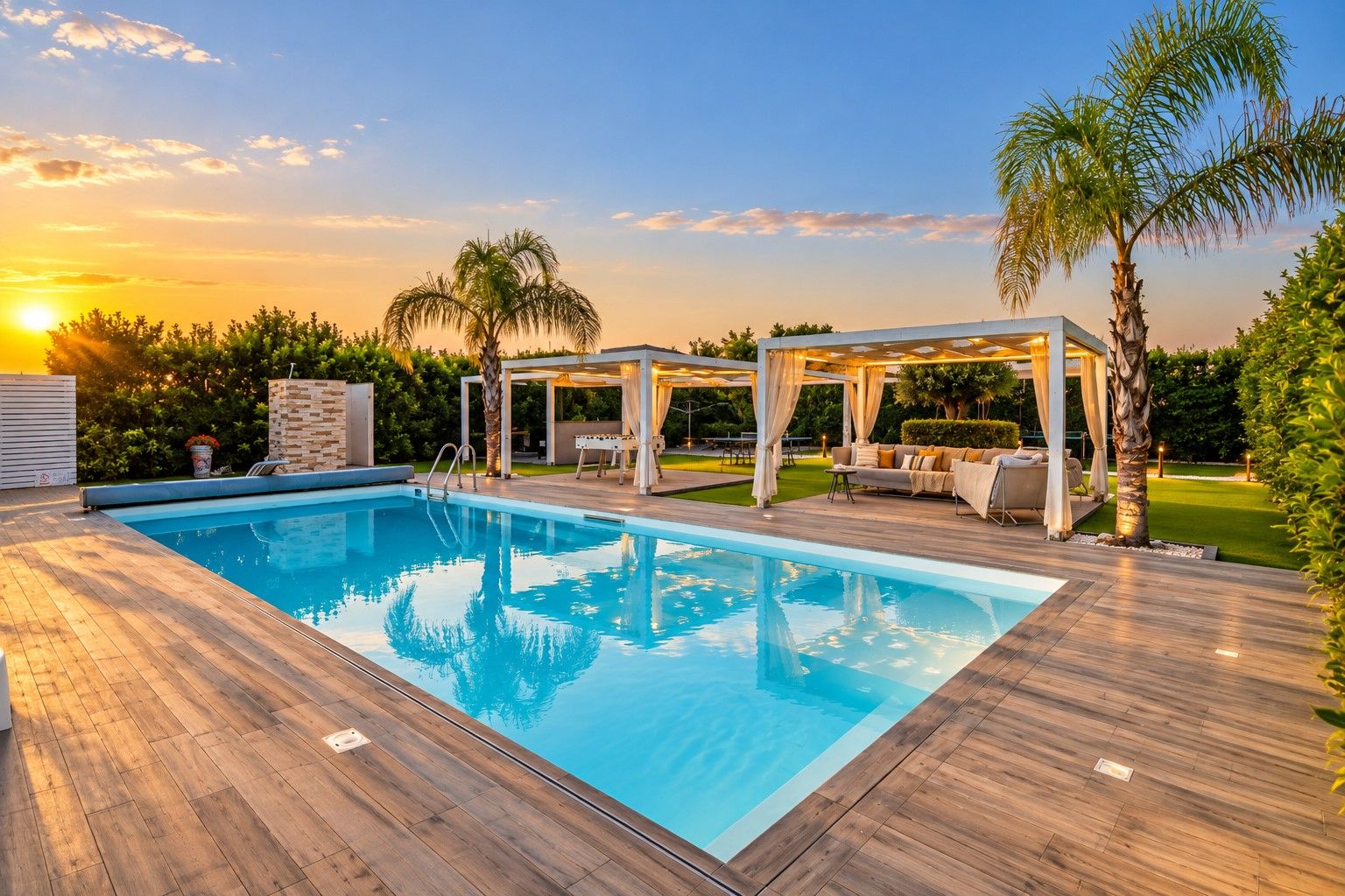 Una piscina rectangular con cubierta de madera, rodeada de mobiliario exterior y una zona techada con cortinas.