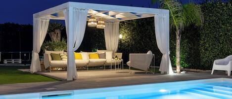 A poolside area with a covered seating area, a pool, and outdoor furniture.