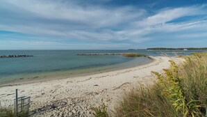 Une plage de sable fin bordée d’une eau claire, un brise-lames rocheux et une végétation herbeuse.