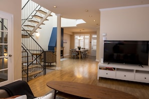 A spacious living area with a wooden floor, a large flat-screen TV, a dining table with chairs, and a staircase with a modern design.
