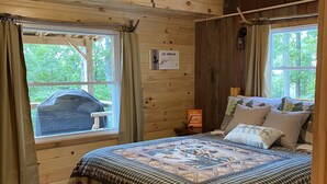 A cozy cabin bedroom with a wooden ceiling, a ceiling fan, a bed with a quilt, a window with curtains, and a sign on the wall. - 🤩 Charming Lake Side Retreat perched above scenic Faubush Creek.  (Nancy)