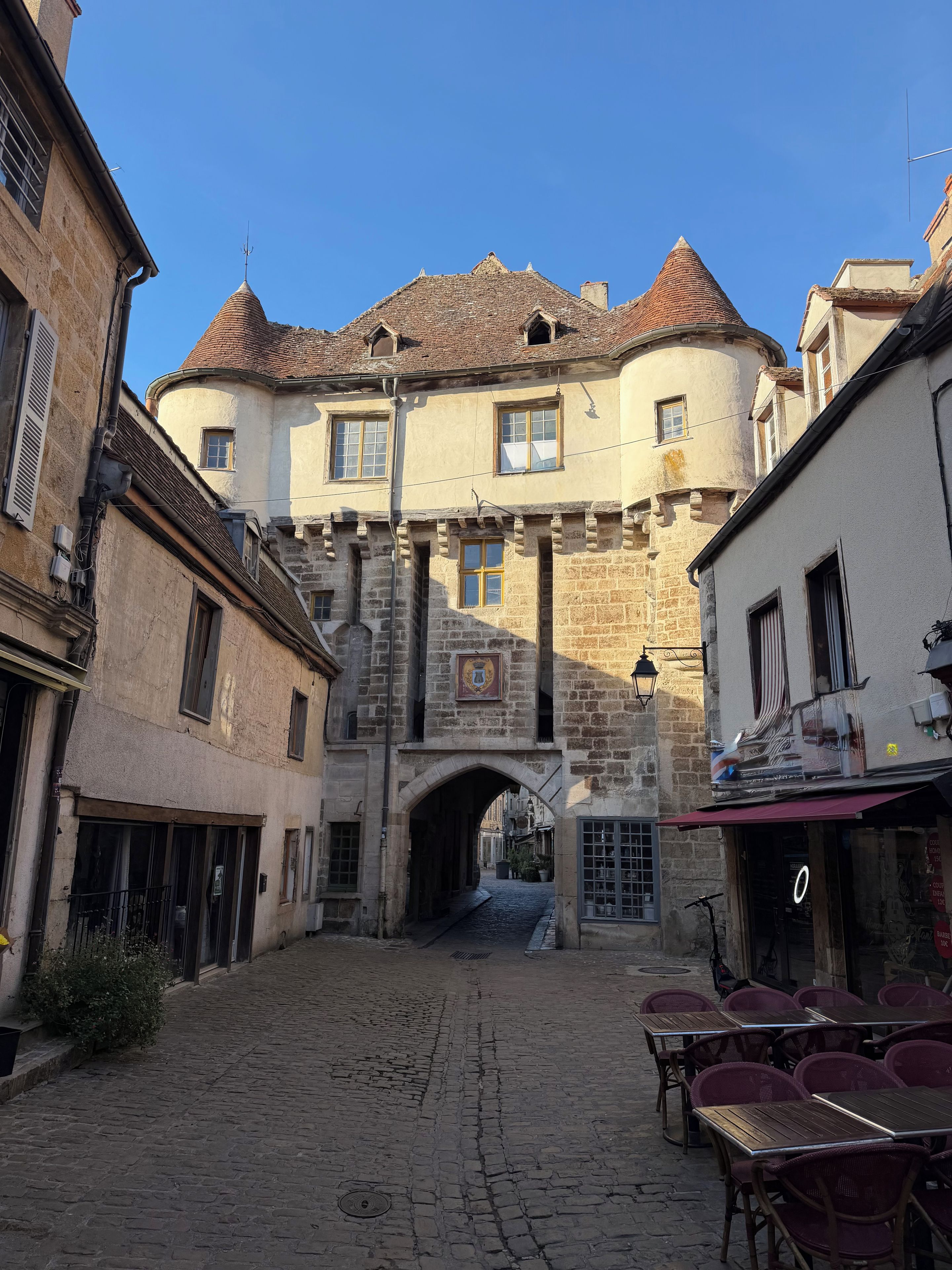 A cobblestone street leading to a historic building with a stone archway, a courtyard with tables and chairs, and a clear blue sky.