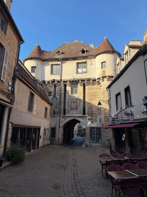 A cobblestone street leading to a historic building with a stone archway, a courtyard with tables and chairs, and a clear blue sky. - Former wine merchant's house and barn, sleeps 14, private pool, stunning views. (Lantilly)
