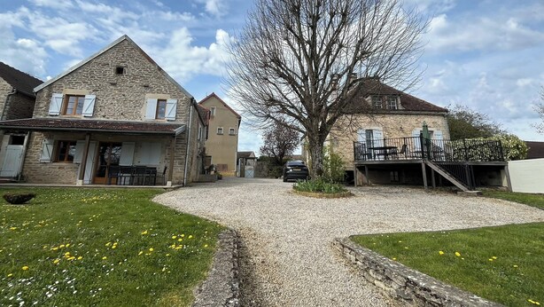 A stone building with a gravel driveway, a tree in the center, and a car parked nearby.