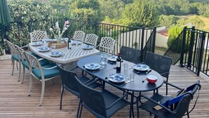 An outdoor dining area with a table set for four, surrounded by chairs, on a wooden deck with a view of a forested landscape.