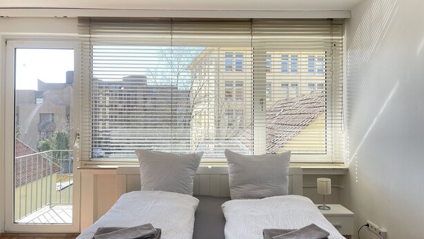 Une chambre moderne avec deux lits, une grande fenêtre avec stores, une vue sur les immeubles et un balcon.