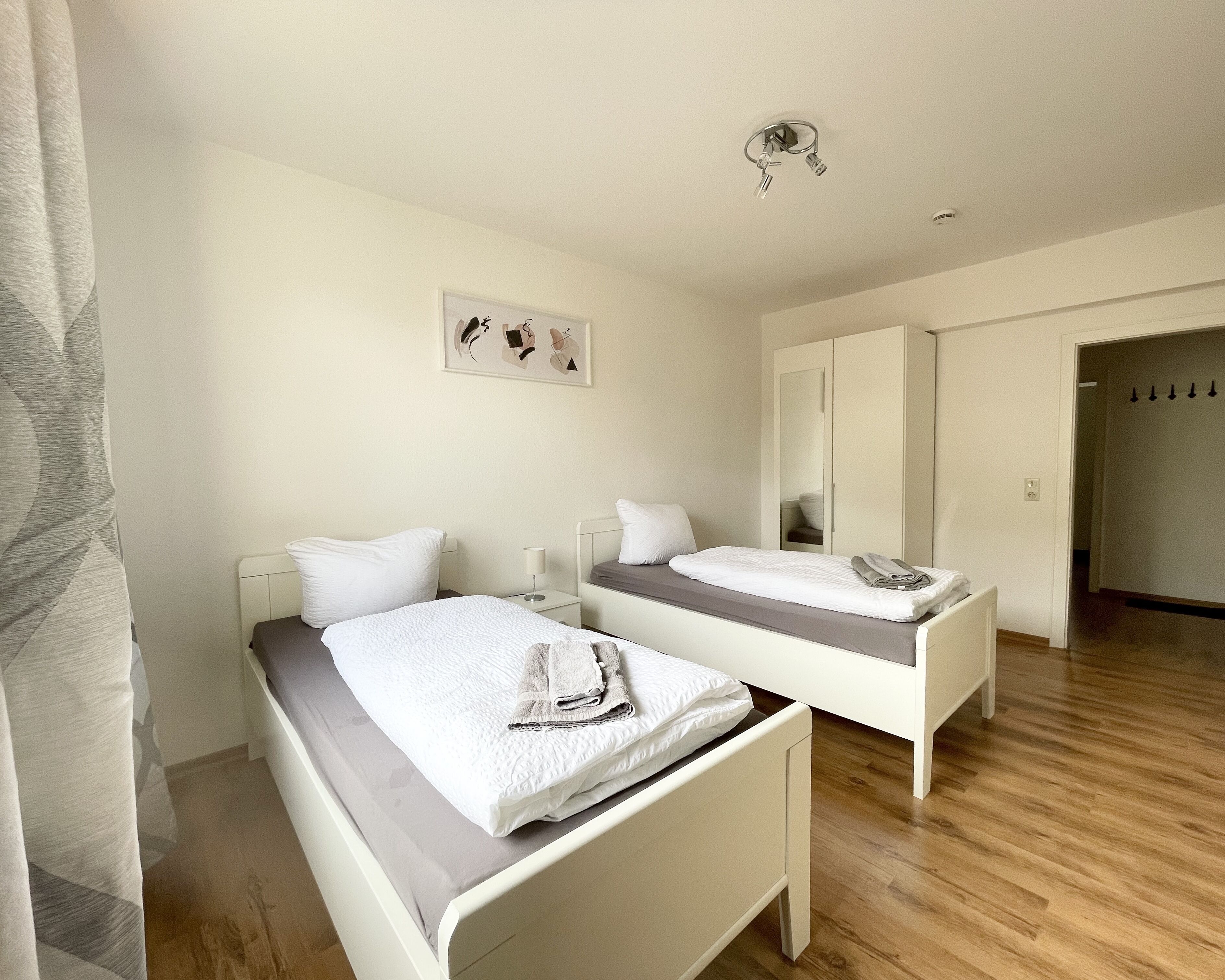 Une chambre d’hôtel avec deux lits simples, une table de chevet avec une lampe, un miroir et une porte donnant sur une autre pièce.