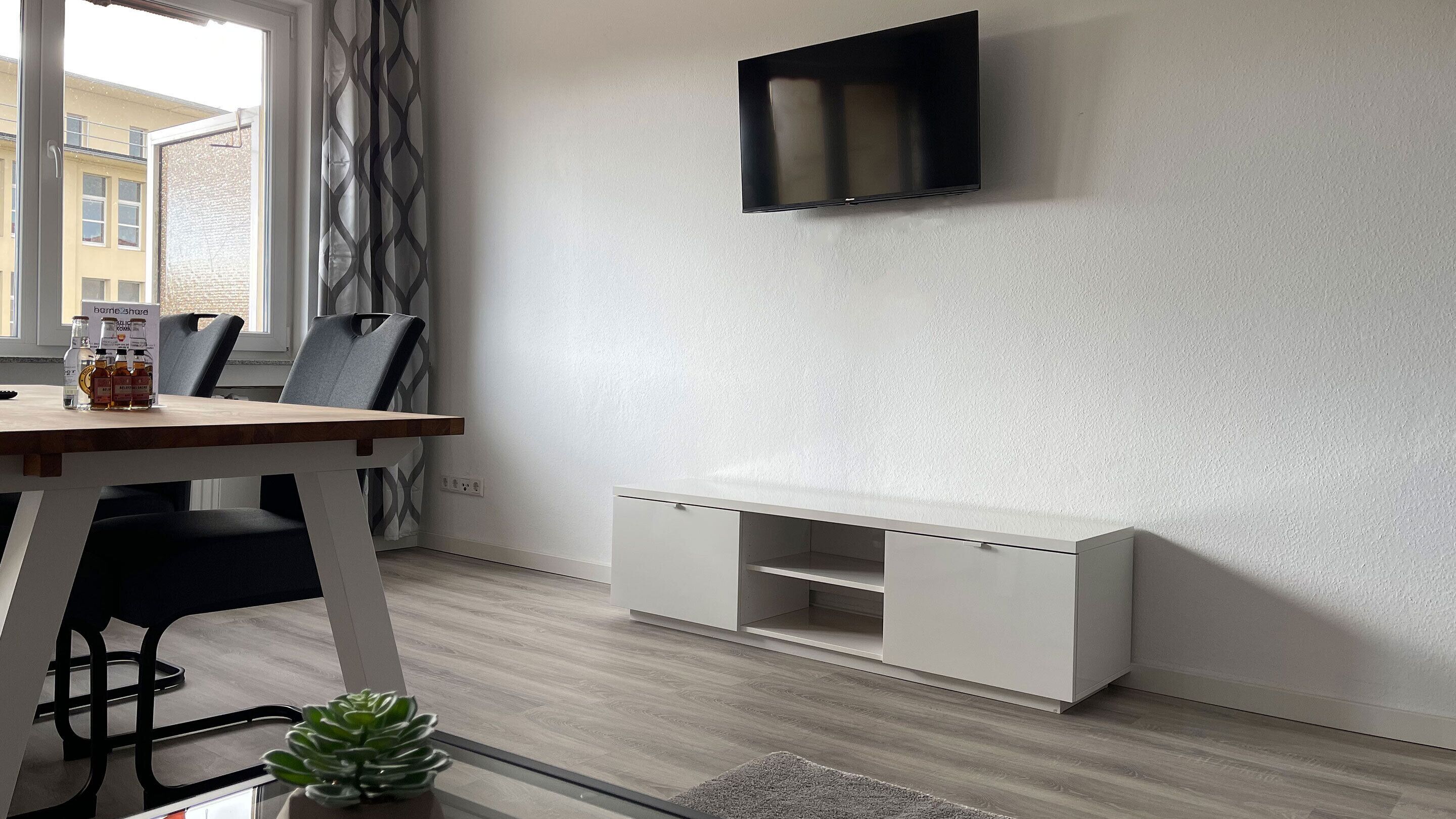 Una sala moderna con un televisor de pantalla plana montado en la pared, una mesa de comedor de madera con sillas, un armario blanco y una pequeña planta en maceta.