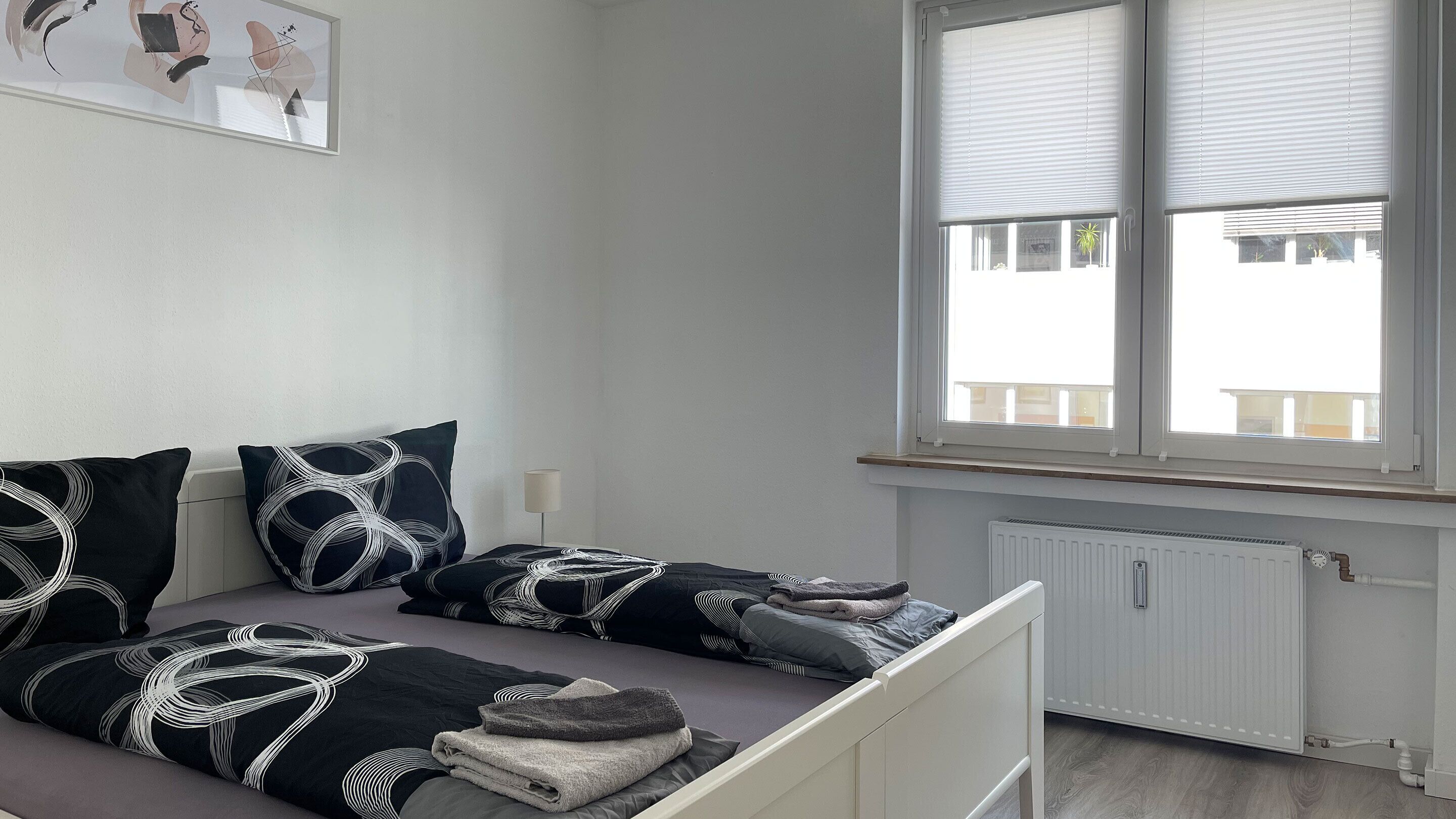Un dormitorio con cama, almohadas, una mesita de noche, lámpara y ventana con persianas.