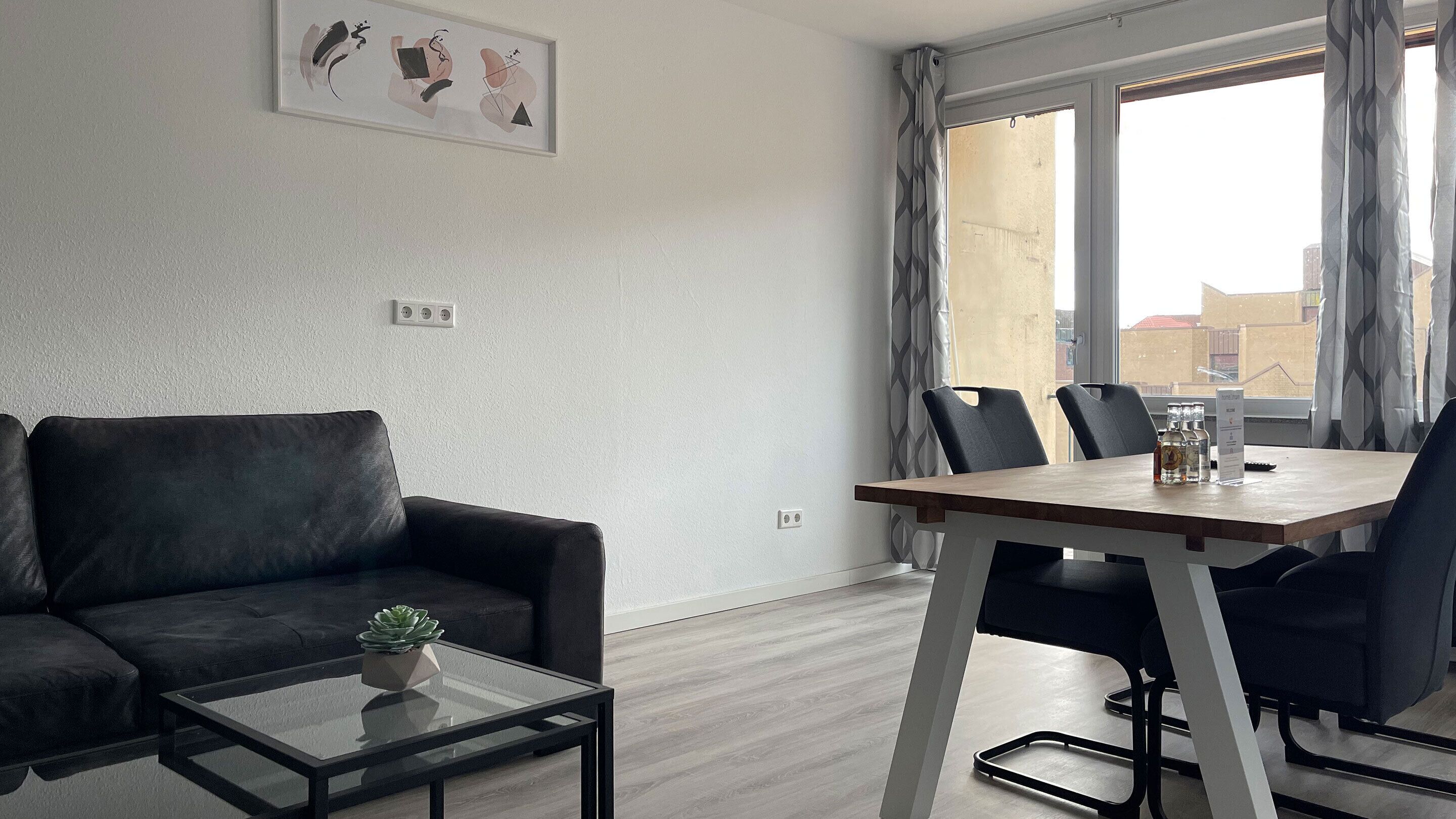 Ein modernes Wohnzimmer mit einer Couch, einem Glastisch und einem Esstisch mit Stühlen. Ein Fenster mit Vorhängen, eine Wandsteckdose und ein gerahmtes Bild über der Couch sind zu sehen.