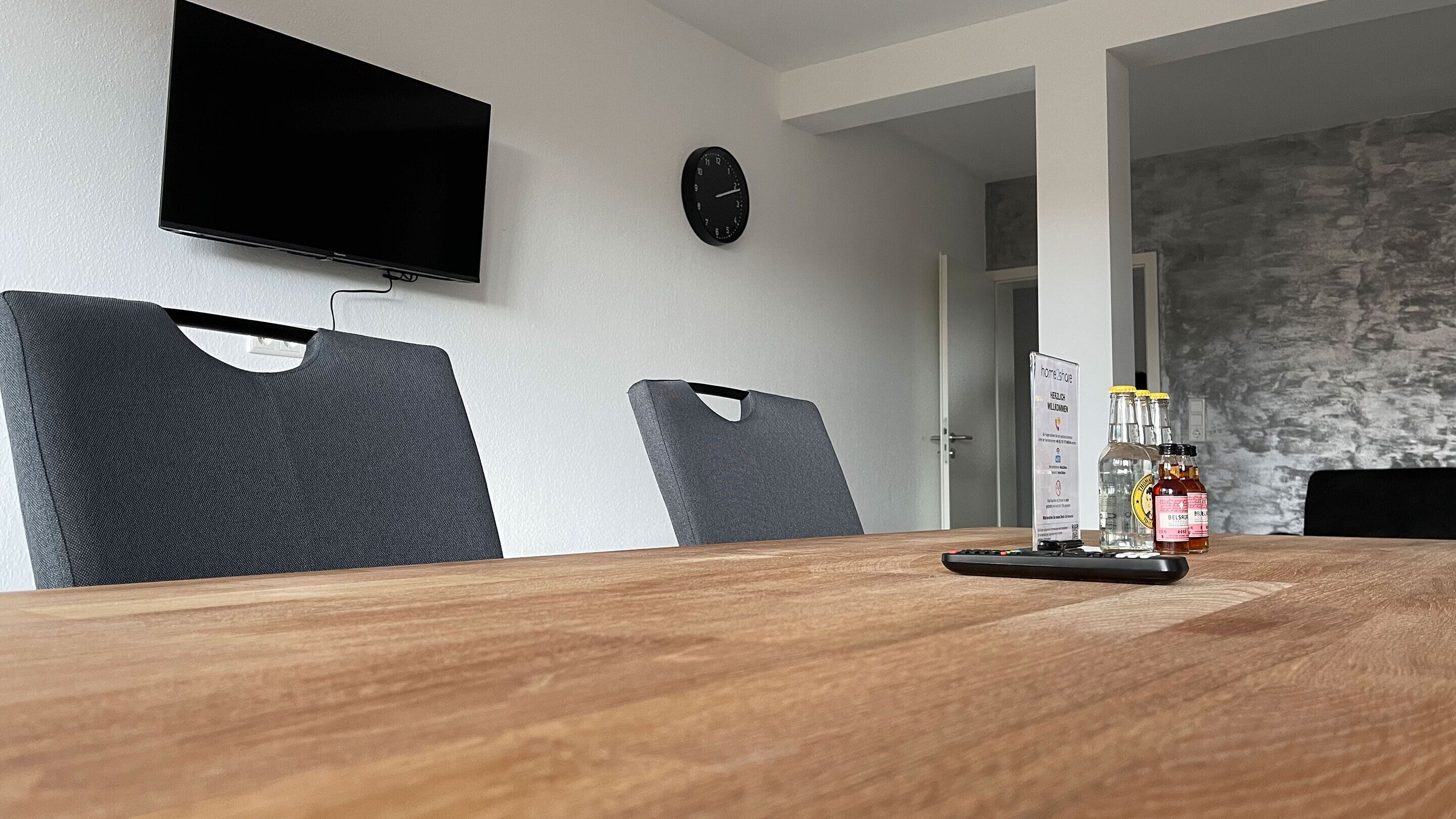 Uma sala de conferências com uma mesa de madeira, duas cadeiras cinzentas, uma TV de tela plana na parede e um relógio.