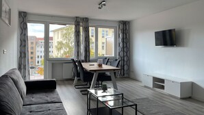 Un salon moderne comprenant un canapé, une table basse en verre, une table à manger avec des chaises, un téléviseur fixé au mur et de grandes fenêtres avec des rideaux.