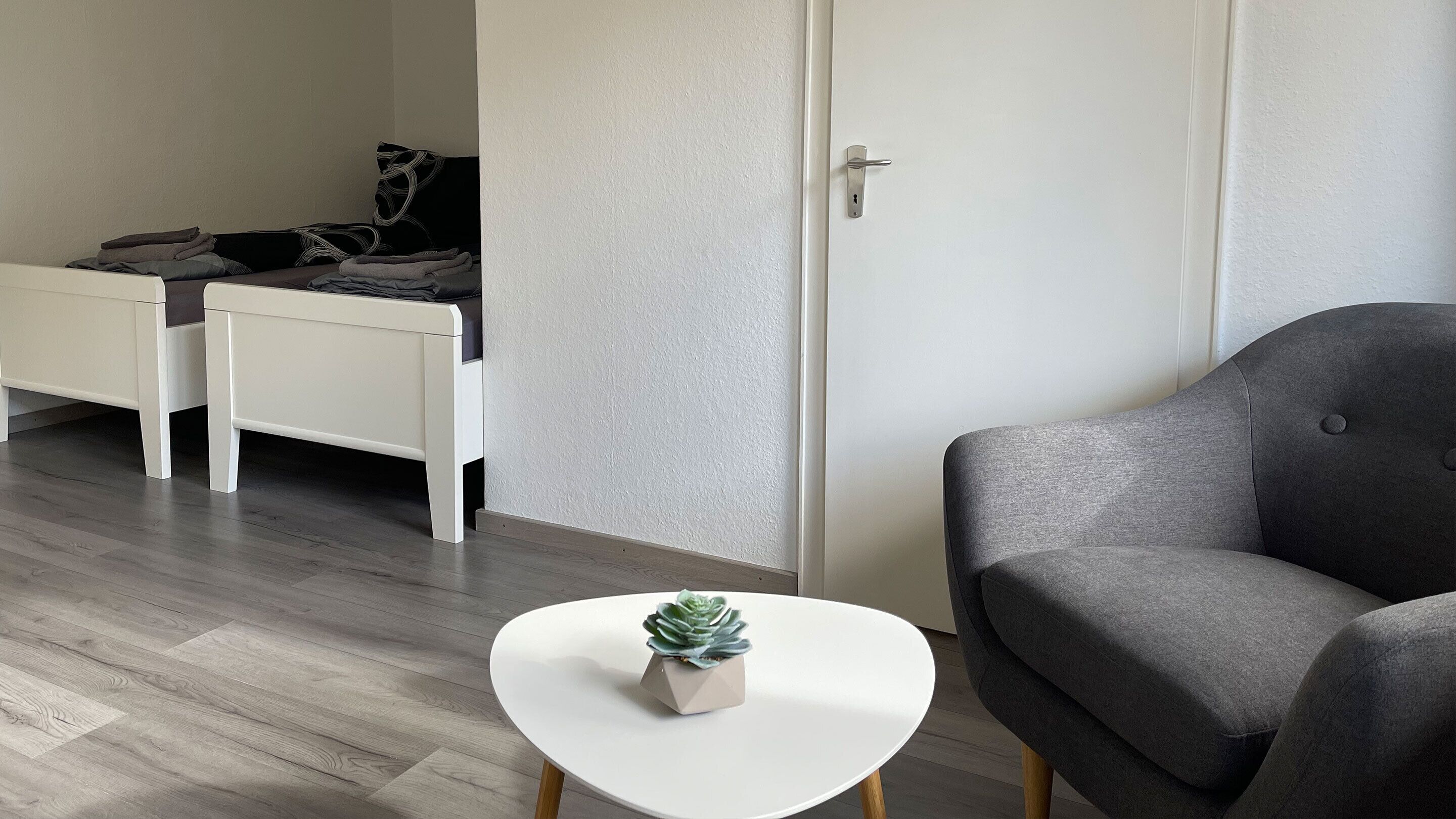 Una habitación moderna con una cama, un escritorio, un sillón gris y una pequeña mesa blanca con una planta suculenta.