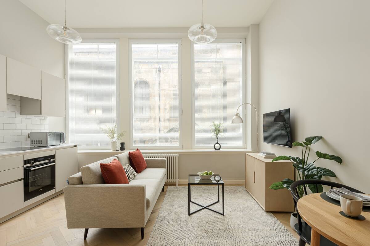 Eine moderne Küche mit weißen Schränken, einem Einbauofen und einer Mikrowelle. Es gibt ein beiges Sofa mit roten Kissen, einen kleinen Tisch mit einer Obstschale und einen Blumentopf.