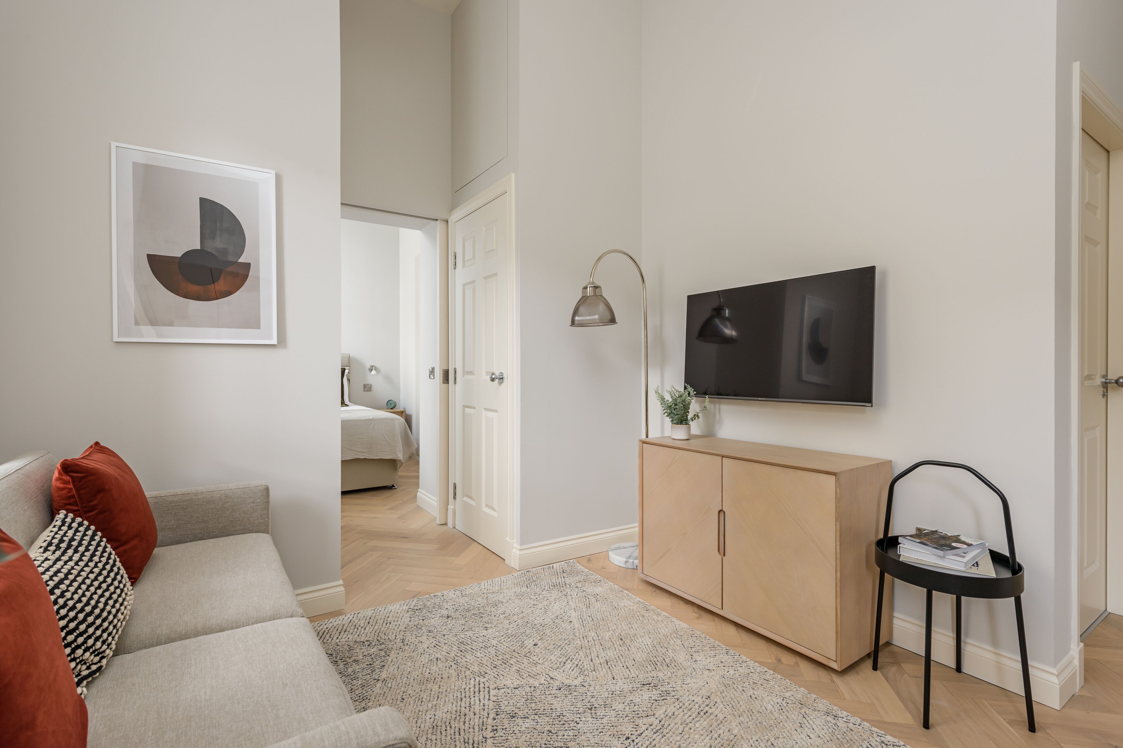 A modern living room with a sofa, a flat-screen TV mounted on the wall, a wooden cabinet, and a small side table with a plant.
