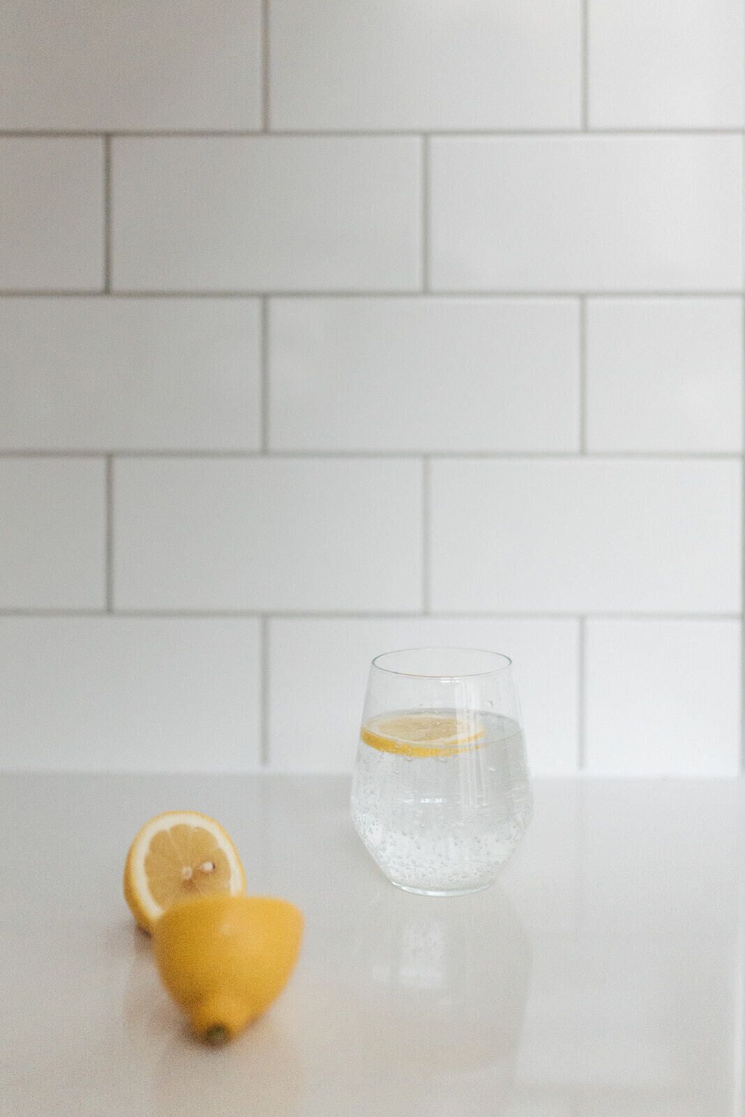 A glass of water with a lemon slice on a white countertop against a white tiled wall.