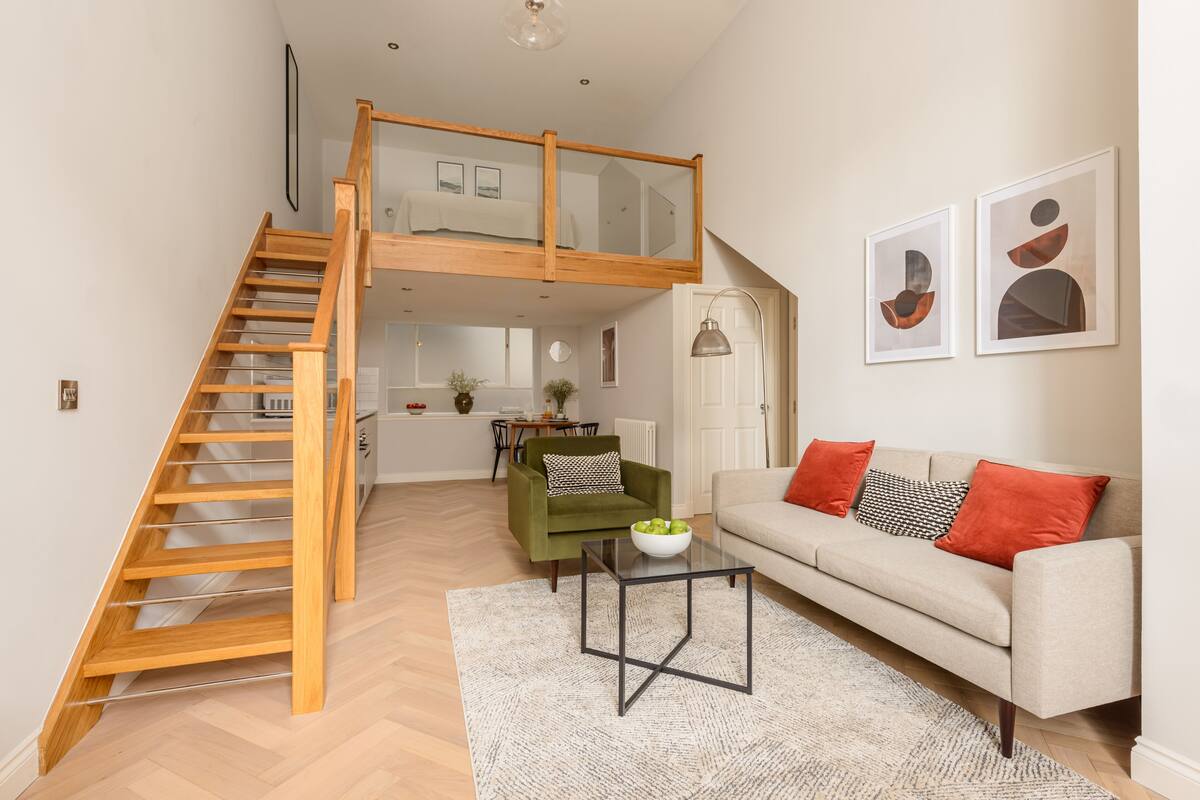 Ein geräumiges Wohnzimmer mit einer Holztreppe zu einem Schlafzimmer im oberen Bereich, ein grüner Sessel, ein beiges Sofa und abstrakte Wandkunst.