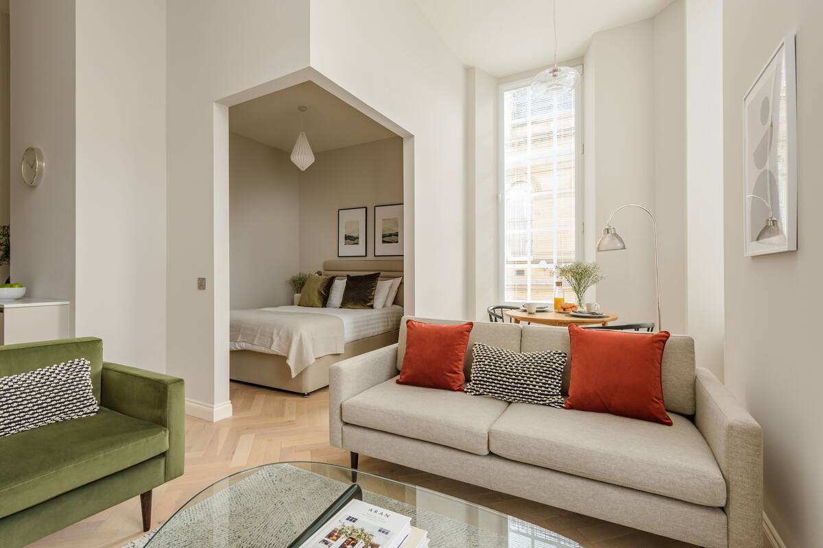 Ein modernes Wohnzimmer mit einer Couch, einem grünen Sessel und einem Glastisch. Im Hintergrund ist ein Bett mit weißem Kopfteil zu sehen.
