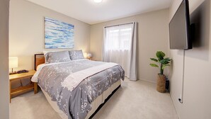Un dormitorio con cama, mesita de noche, lámpara, una obra de arte, ventana, cortinas y una planta.