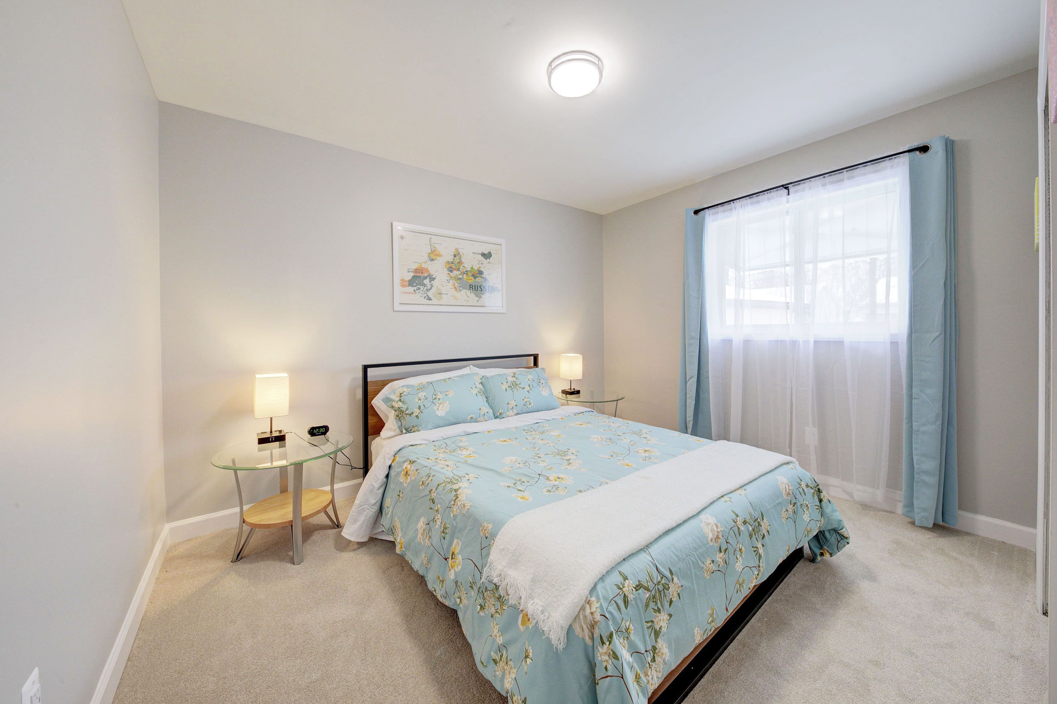 A bedroom with a bed, bedside tables, a lamp, a map on the wall, and a window with curtains.