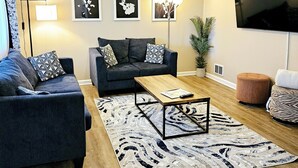 Una sala moderna con un sofá gris oscuro, una mesa de centro de madera y un tapete estampado. Se observan cuadros enmarcados en la pared, una lámpara de pie y un televisor.