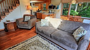 Ein Wohnzimmer mit einer Couch, Sesseln, Holzboden, Teppich und einer Treppe, im Hintergrund ist eine Küche zu sehen.