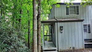 Ein kleines, eingeschossiges Holzhaus mit Veranda, umgeben von dichtem Grünwerk.