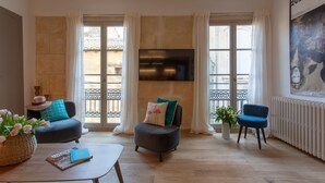 A living room with a sofa, armchair, and a coffee table. There is a TV mounted on the wall, a poster on the wall, and a window with curtains. - Flamingo Duplex, historical centre, l'Hauture, Arles (Arles)