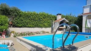 Ein Schwimmbecken mit einer Metallleiter, umgeben von einer gefliesten Terrasse und einem Hecken.