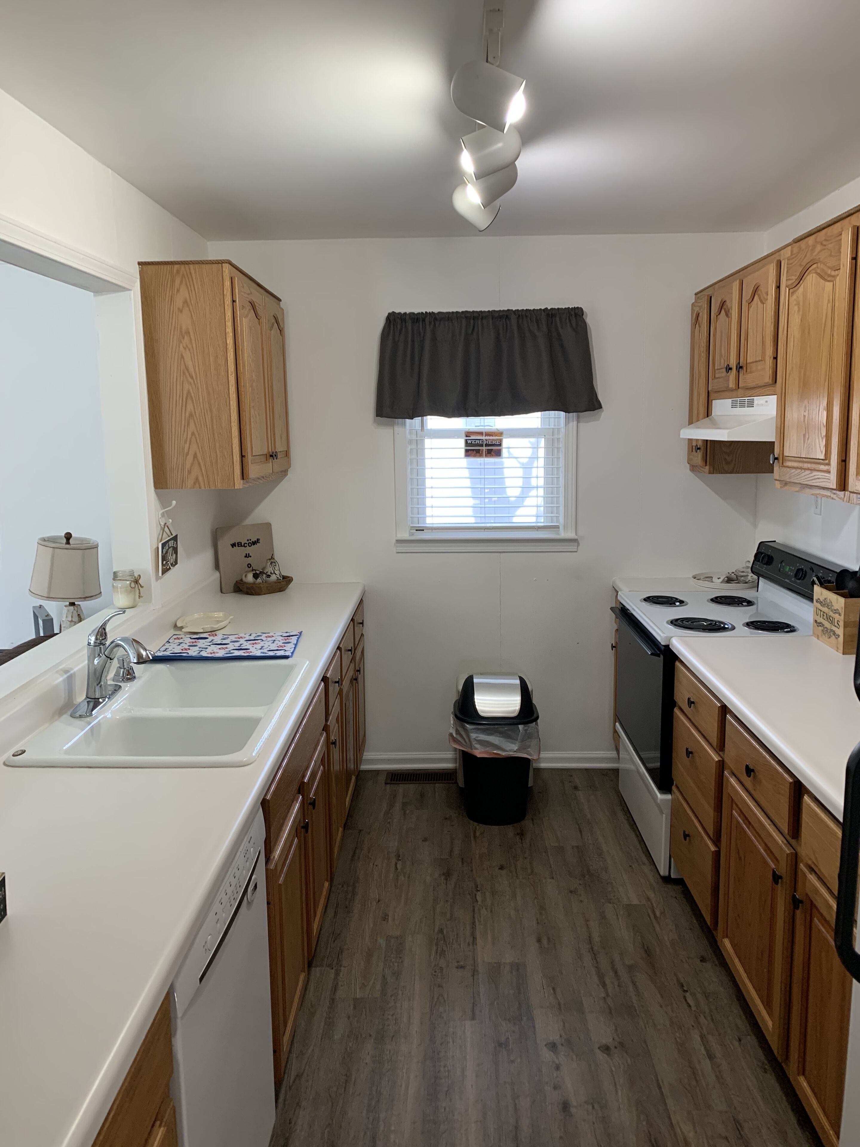 A kitchen with wooden cabinets, a white countertop, a sink, a stove, and a trash can.