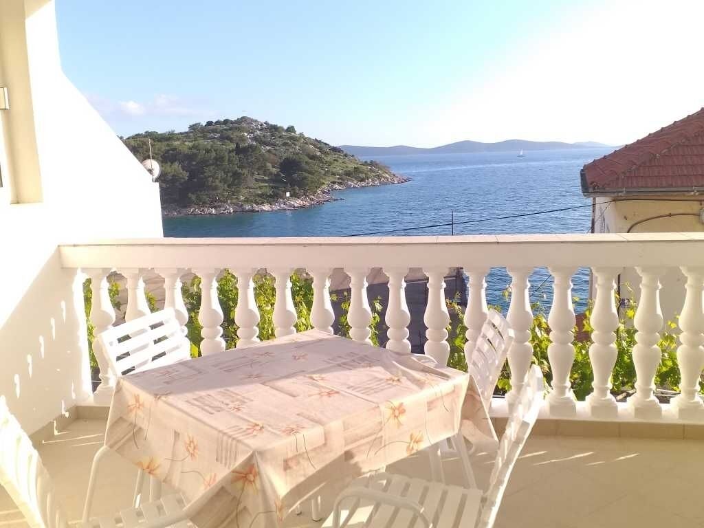 A balcony with a table and chairs, overlooking a body of water and a green hillside.