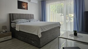 Quarto moderno com cama cinza, roupa de cama branca, ar condicionado fixado na parede, mesa de cabeceira com abajur e uma grande janela com cortinas azuis.