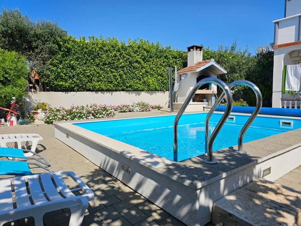 Une piscine avec un escalier en métal, entourée d’une terrasse carrelée et d’une haie.