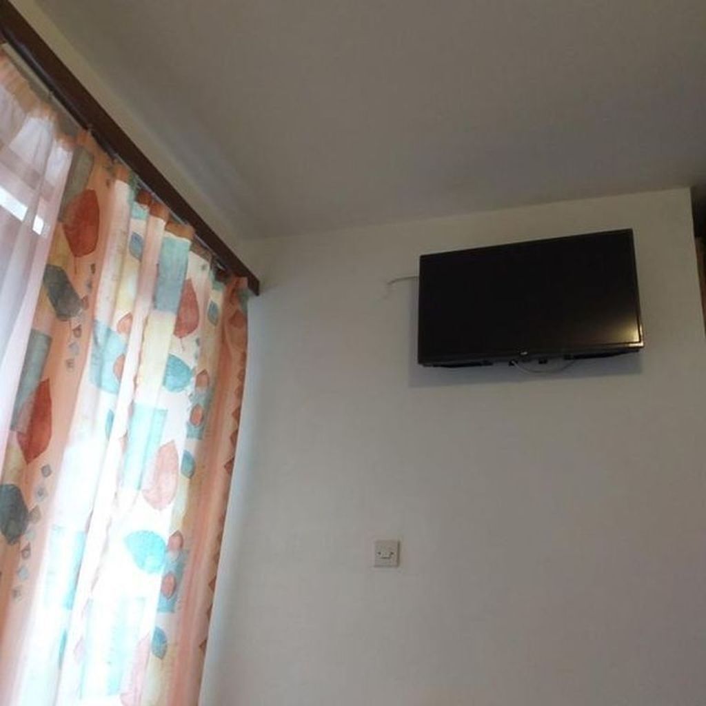 Ein Zimmer mit an der Wand befestigtem Fernseher, einer Steckdose und einem Fenster mit Vorhang.