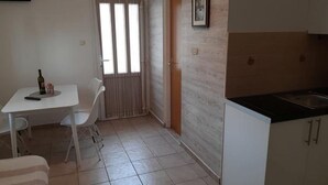 Et lille, rent værelse med tekøkken, spiseplads med bord og stole samt et vindue med gardiner.