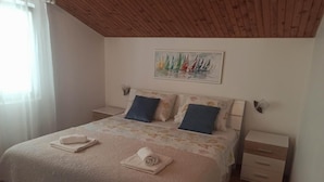 Une chambre avec un plafond en bois, un lit recouvert d’une couvre-lit à motifs floraux, deux oreillers, une table de chevet et un tableau représentant des voiliers accroché au mur.
