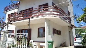 Un immeuble de deux étages doté d’un balcon, d’une poubelle et d’une voiture garée.
