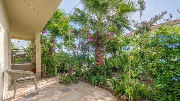 A patio area with stone flooring, a wicker chair, and lush greenery including palm trees and flowering plants.