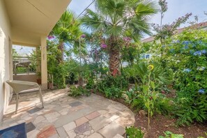 Eine Terrassenfläche mit Steinboden, einem Korbstuhl und üppiger Bepflanzung, darunter Palmen und blühende Pflanzen.