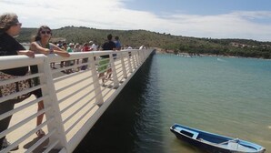 A pier with people walking along it, overlooking a calm body of water and a hilly landscape in the distance.