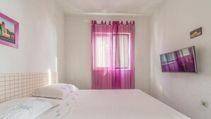 A bedroom with a bed, a window with pink curtains, a television on the wall, and a painting on the wall.