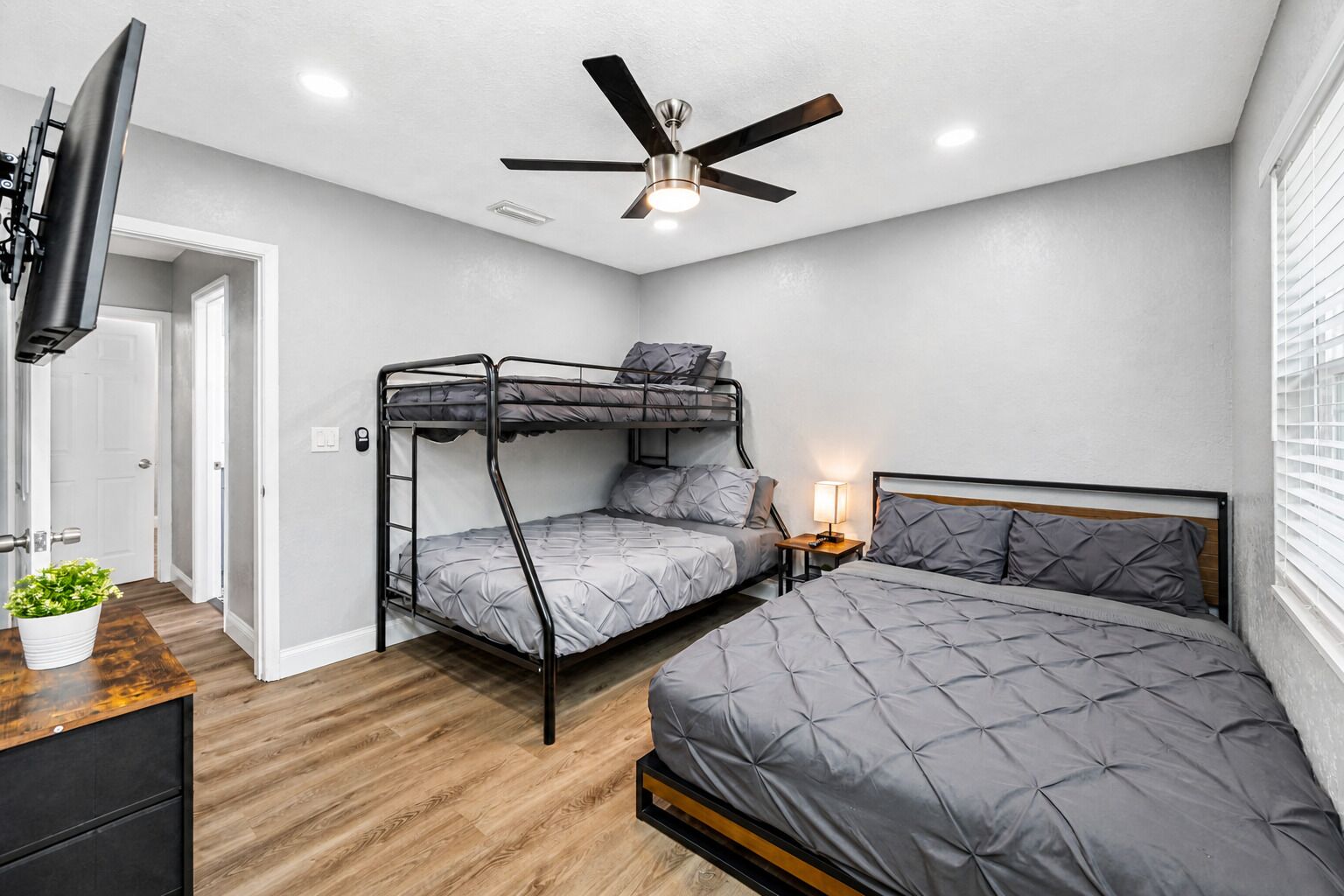 Un dormitorio con una cama litera, una cama individual, un ventilador de techo, un televisor montado en la pared y una mesita de noche con lámpara.