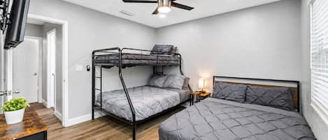 Un dormitorio con una cama litera, una cama individual, un ventilador de techo, un televisor montado en la pared y una mesita de noche con lámpara.