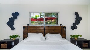 A bedroom with a wooden headboard, a large bed with white bedding, two bedside tables with plants, and a window with a view of greenery and a red umbrella.