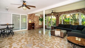 A spacious living area with a large sofa, a coffee table, and a dining table with chairs. There are two ceiling fans, a brick wall, and large windows with a view of greenery.