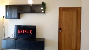 A living room with a black entertainment unit, a TV displaying the Netflix logo, a wooden door, and a modern light fixture.